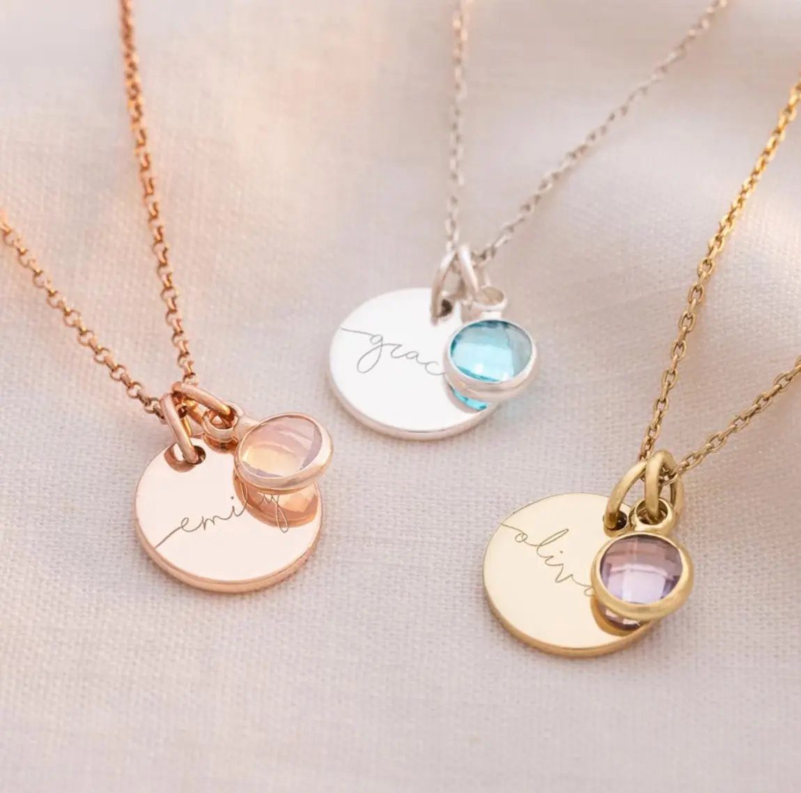 Three personalized necklaces with engraved name discs and colorful gemstones on white fabric.