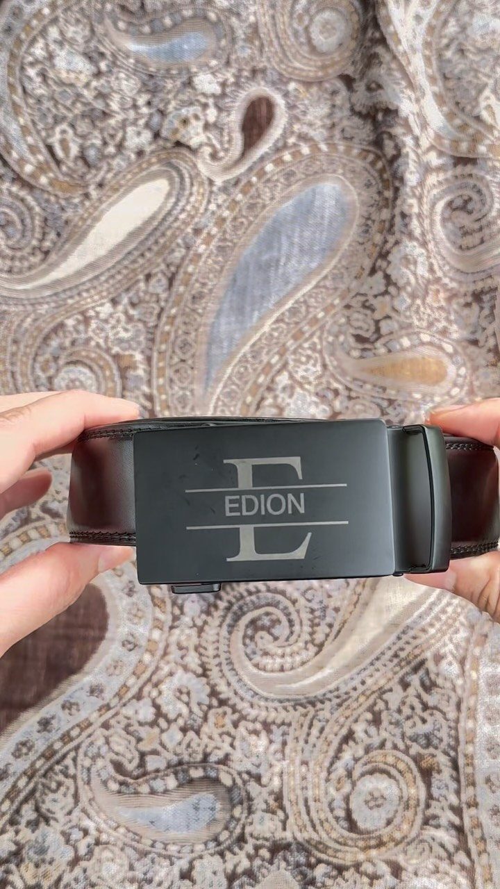 Black leather belt with an engraved buckle held over a paisley patterned rug.