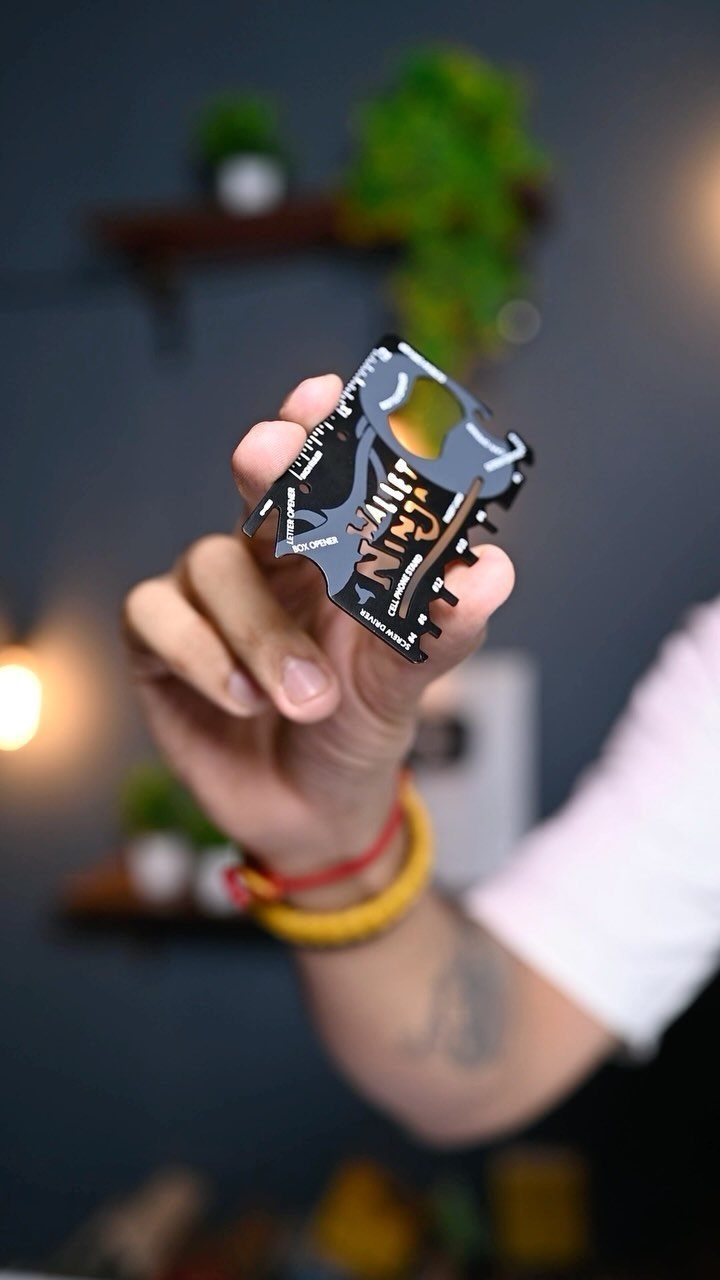 Hand holding a black metal wallet multitool card against a blurred indoor background.