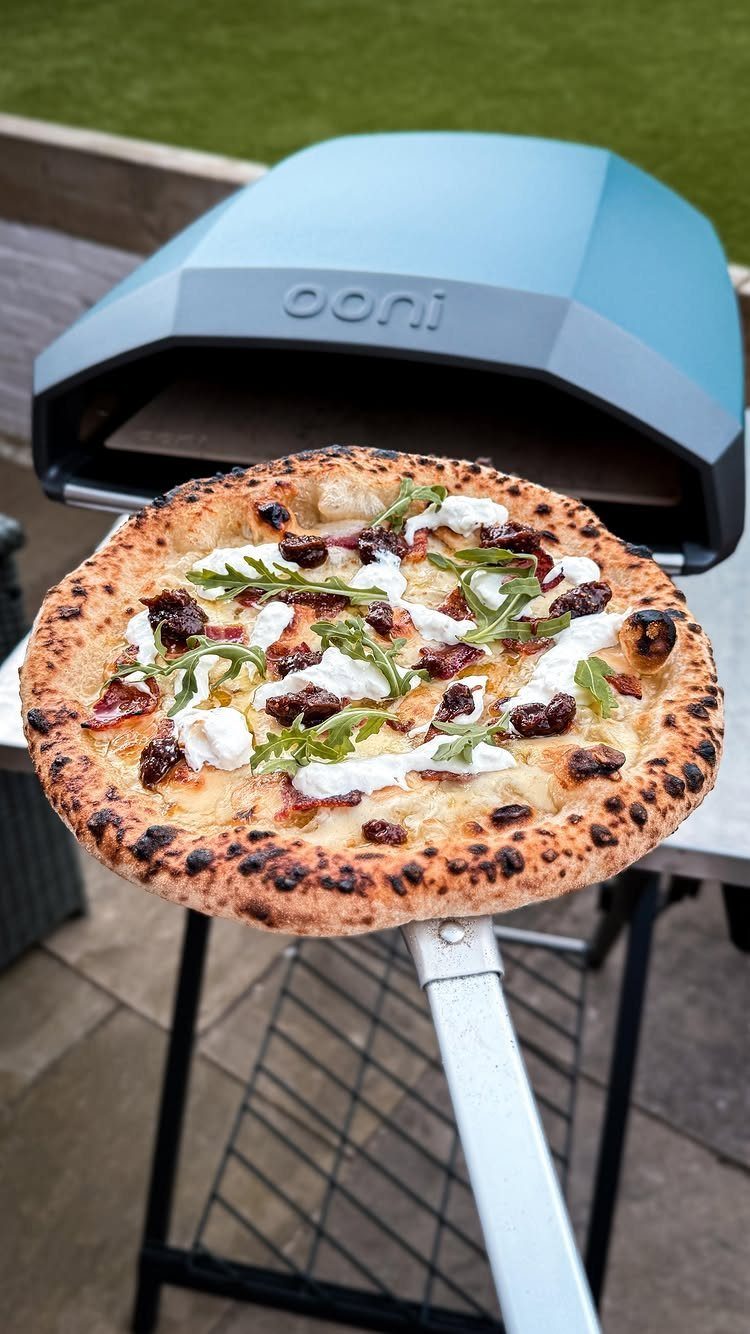 Freshly baked pizza with arugula and cheese in front of a blue Ooni oven.