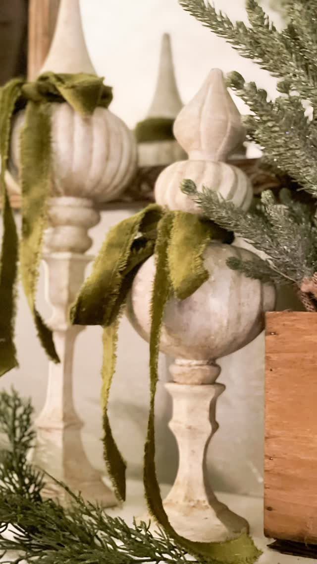 Ornate cream stone finials tied with olive green velvet ribbons among evergreen branches.