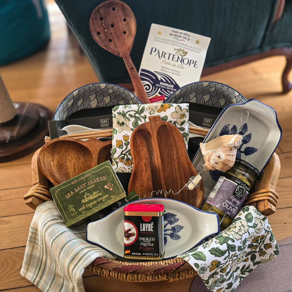Wicker gift basket containing wooden serving tools, ceramic dishes, and gourmet food items.