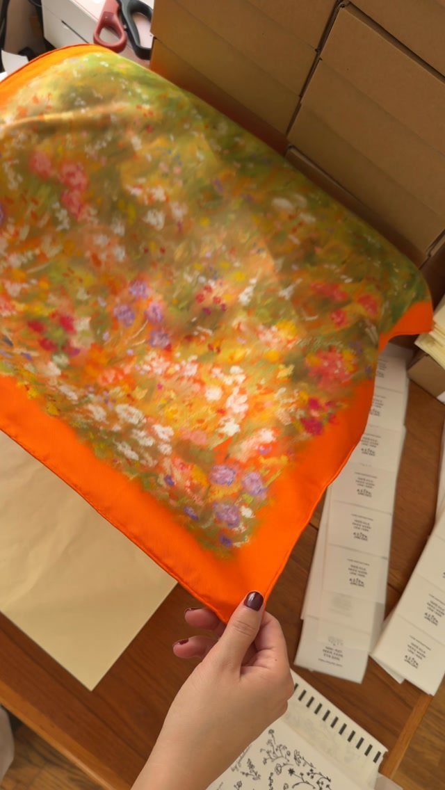 Colorful floral silk scarf with orange border held by a hand over cardboard