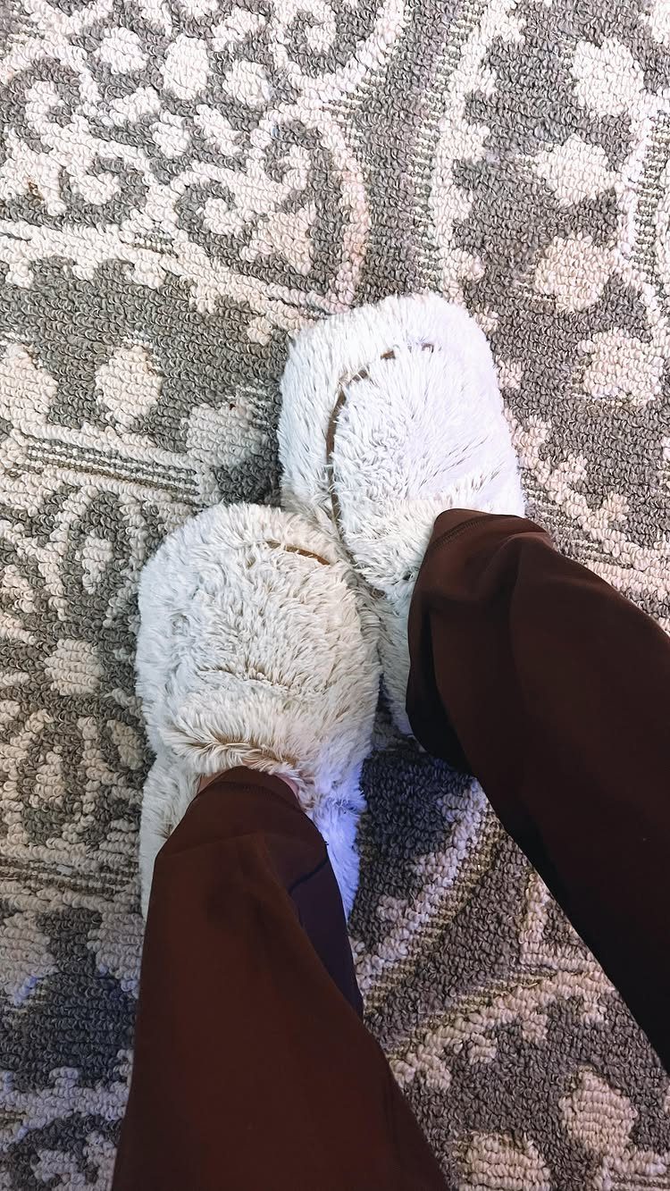 White fuzzy slippers worn by a person standing on a patterned grey rug.