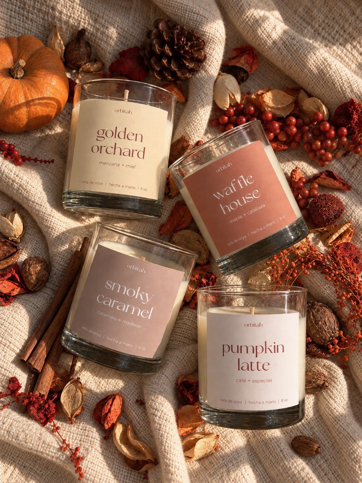 Four autumn-themed scented candles arranged on beige fabric with pumpkins and cinnamon sticks.