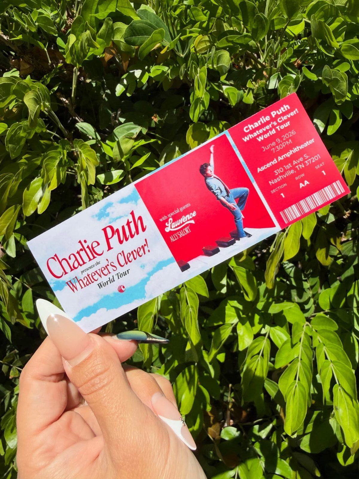 Hand holding a red and white Charlie Puth concert ticket against green leaves.