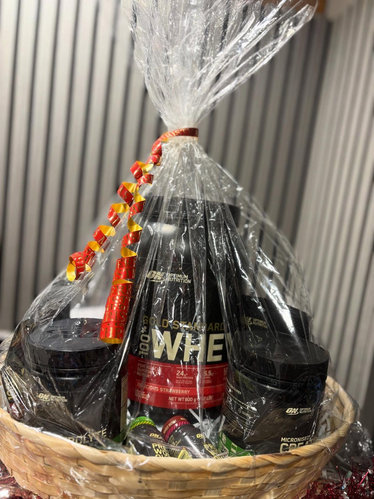 Protein powder tub and supplement jars in a cellophane-wrapped wicker gift basket.