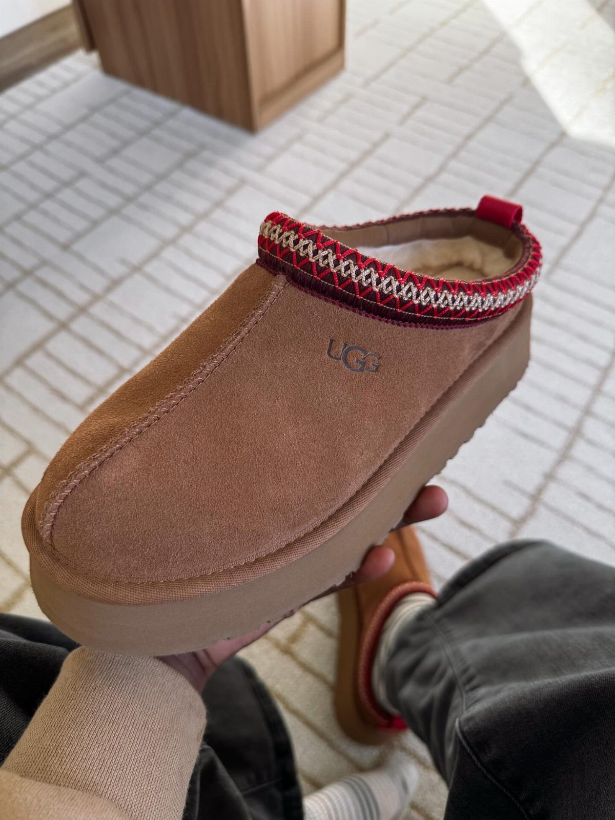 Brown suede Ugg slipper with red patterned trim held by a person.