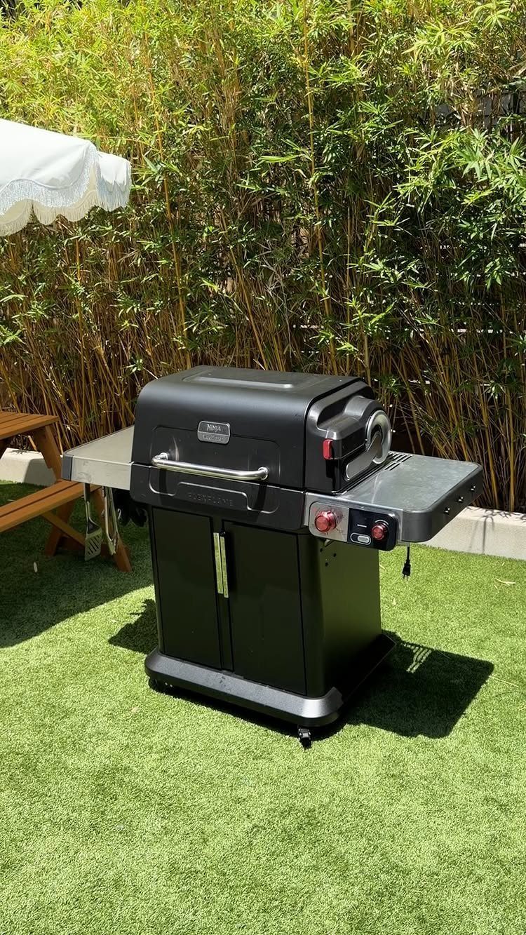 Black outdoor grill on green artificial grass in front of a bamboo hedge.