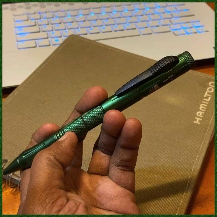 Metallic green tactical pen held in a hand over a leather notebook