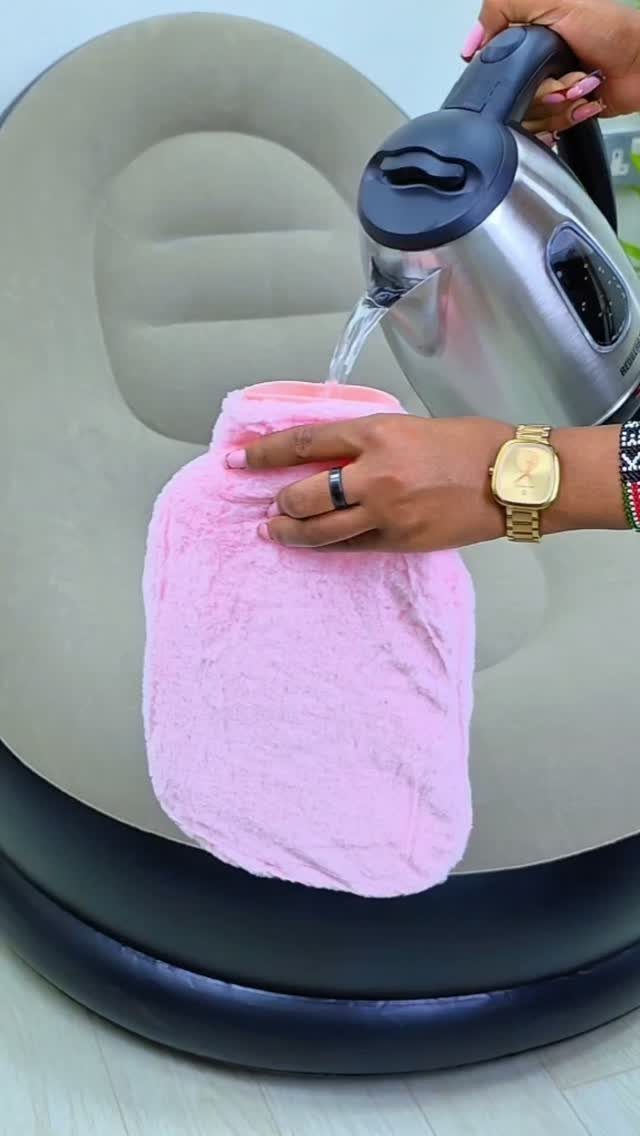 Hands pouring water from a steel kettle into a pink fluffy hot water bottle.