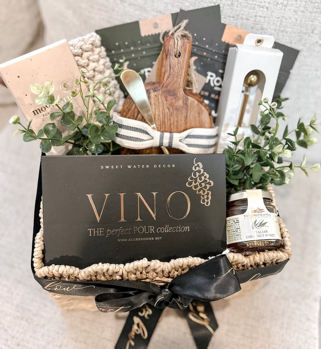 Woven gift basket containing wine accessories, a wooden board, and green foliage.