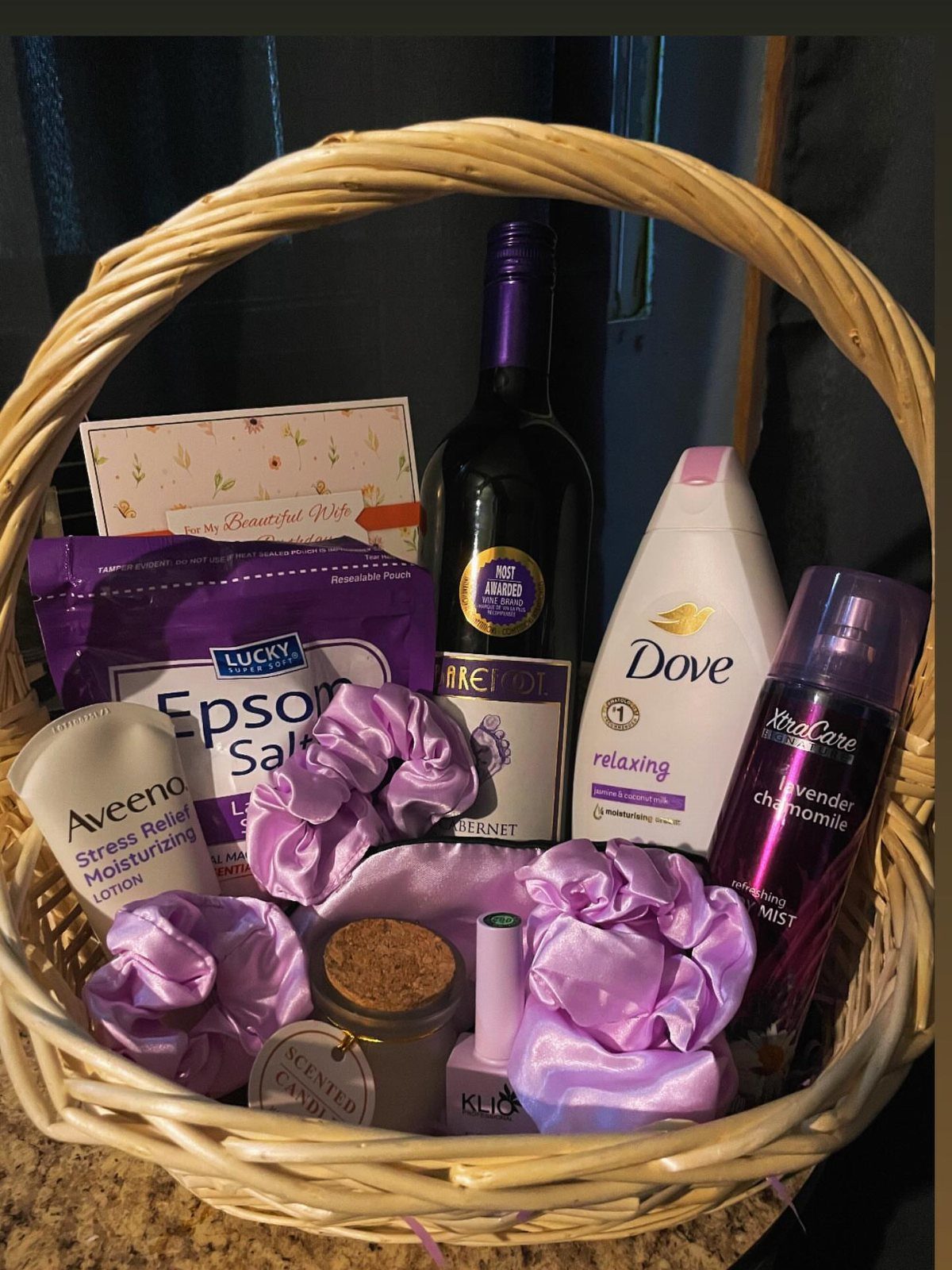Wicker gift basket containing red wine, purple beauty products, and scented candle.